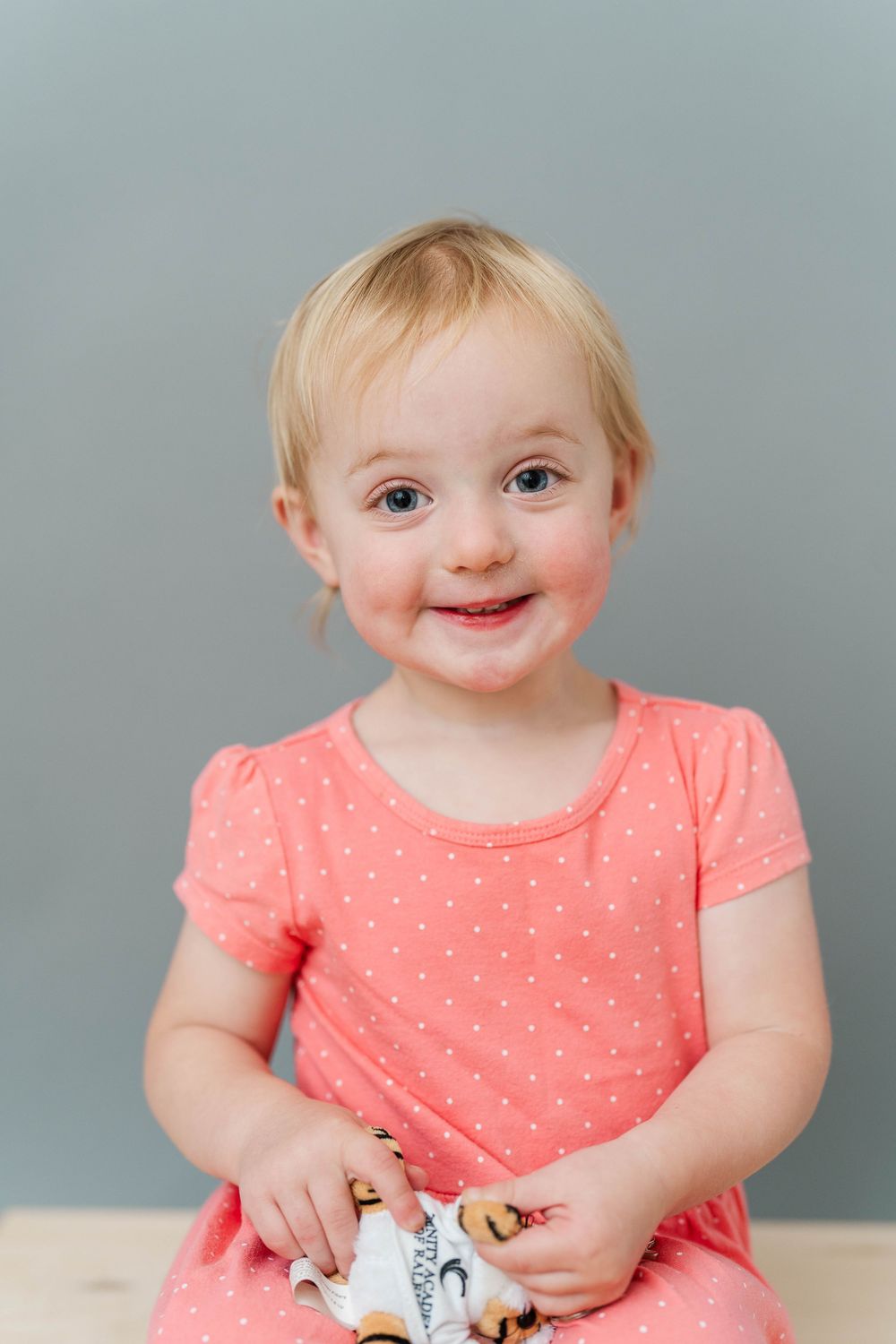 Smiling toddler girl with blonde hair wearing a pink polka dot dress, holding a toy against a gray background.