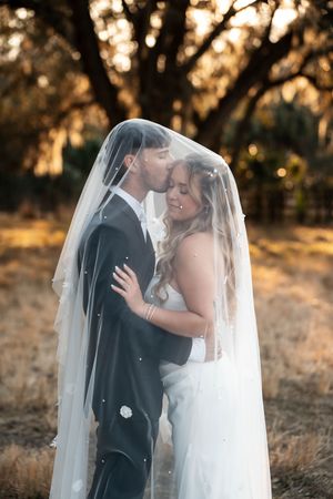 Bride and groom share a romantic kiss under a flowing veil at golden hour in a rustic outdoor setting.