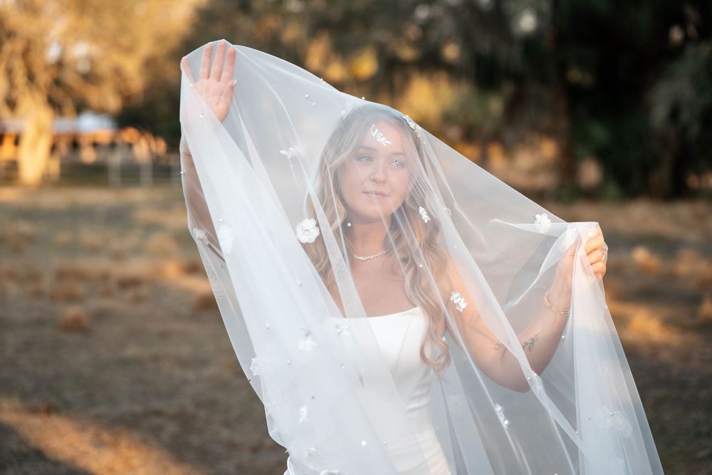 Bride in white dress holding sheer veil with floral details outdoors in golden light.