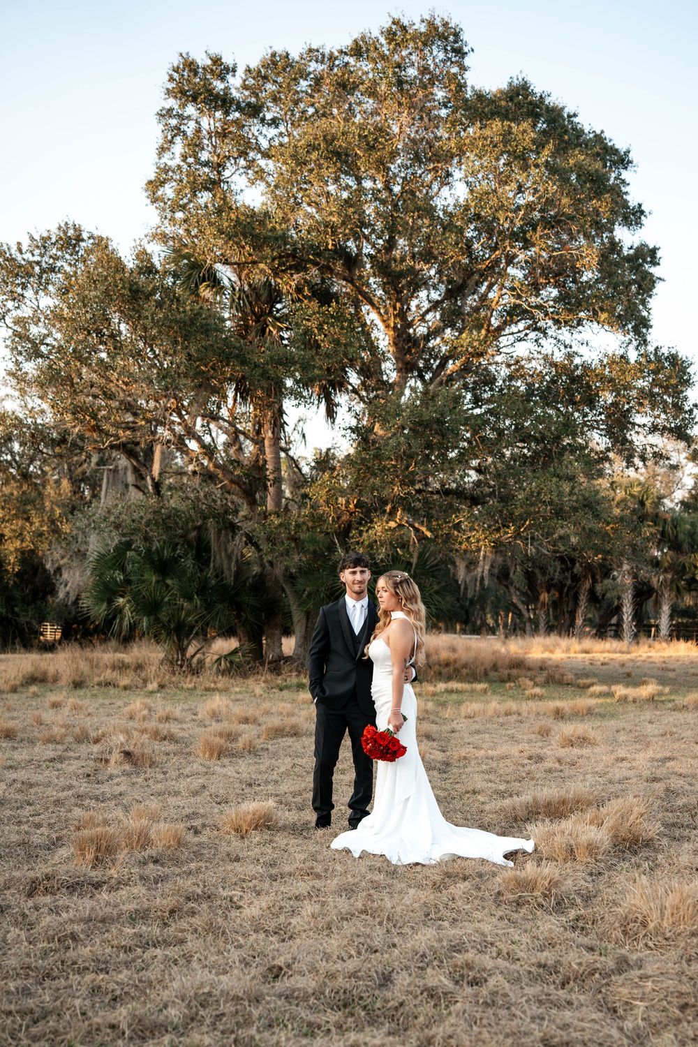 Bride in white gown holding red bouquet stands with groom in black suit in golden field before large oak tree.
