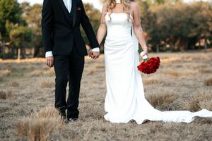 Bride in white gown holding red roses bouquet and groom in black tuxedo holding hands in a dry field.