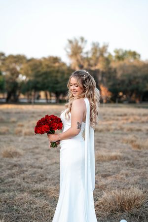 Bride in white gown holding red rose bouquet standing in golden field with trees in background.