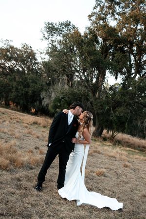 Bride and groom sharing a romantic kiss in a golden field with oak trees, bride in elegant white gown.