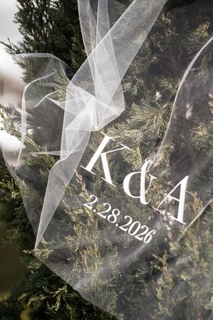 Clear acrylic wedding sign with K&A monogram and date 2.28.2026, adorned with tulle ribbon against evergreen foliage.