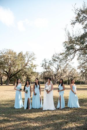 Bride in white gown poses with five bridesmaids in light blue dresses outdoors among oak trees.