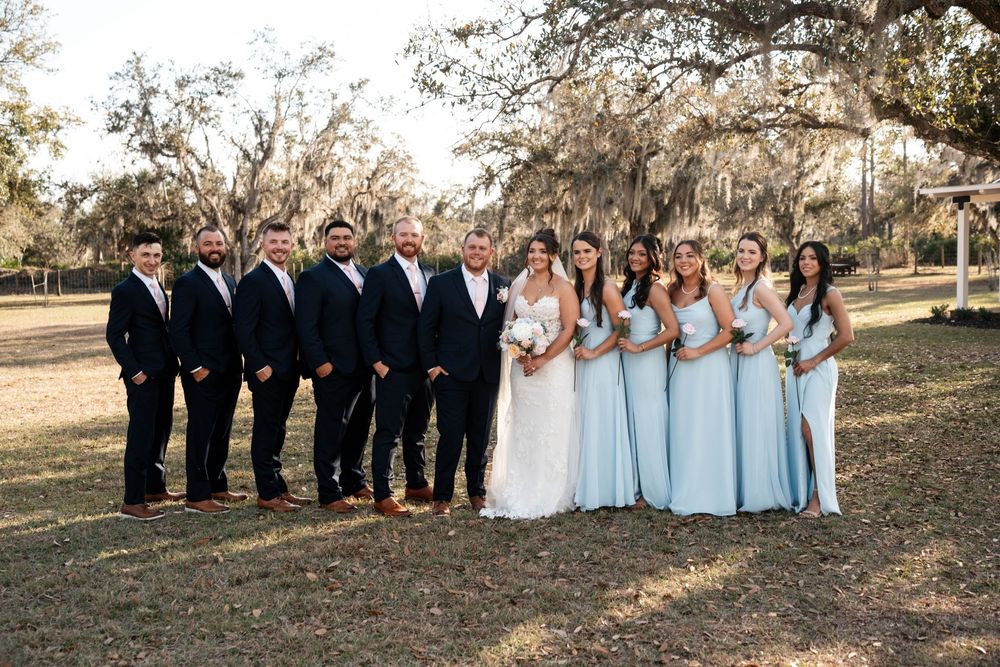 Wedding party poses outdoors, groomsmen in navy suits and bridesmaids in light blue gowns, with bride and groom centered.