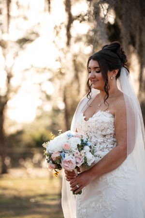Bride in strapless lace gown holds pink and white bouquet while wearing veil outdoors.