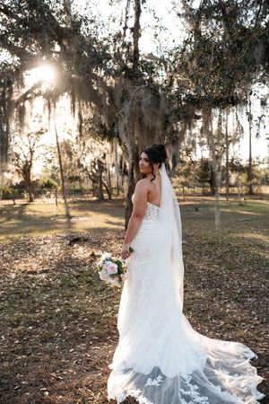 Bride in lace mermaid gown with veil holds bouquet under Spanish moss-draped oak trees in golden sunlight.