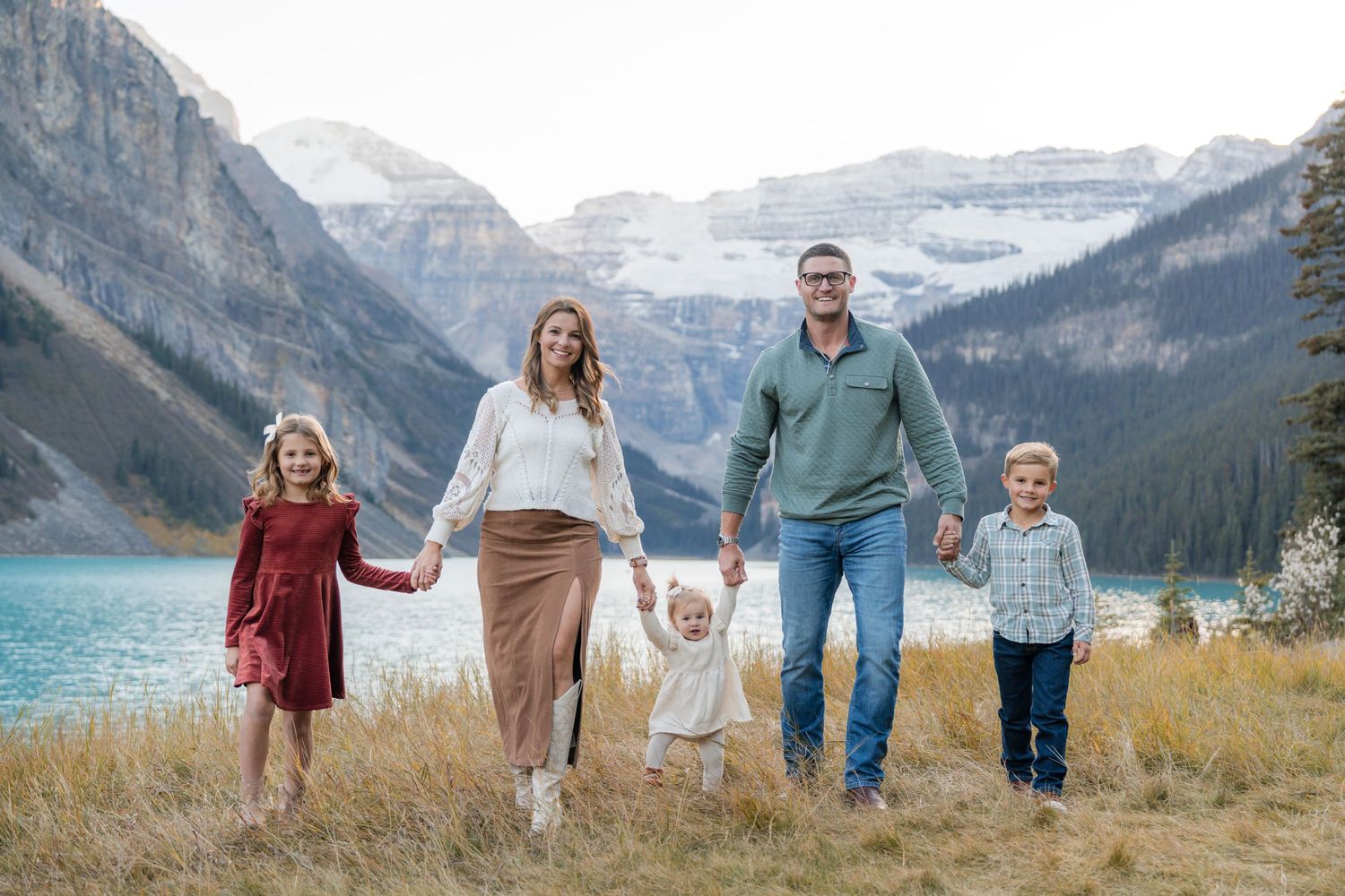 Family of five posing outdoors near turquoise Lake Louise with snow-capped Rocky Mountains in background.