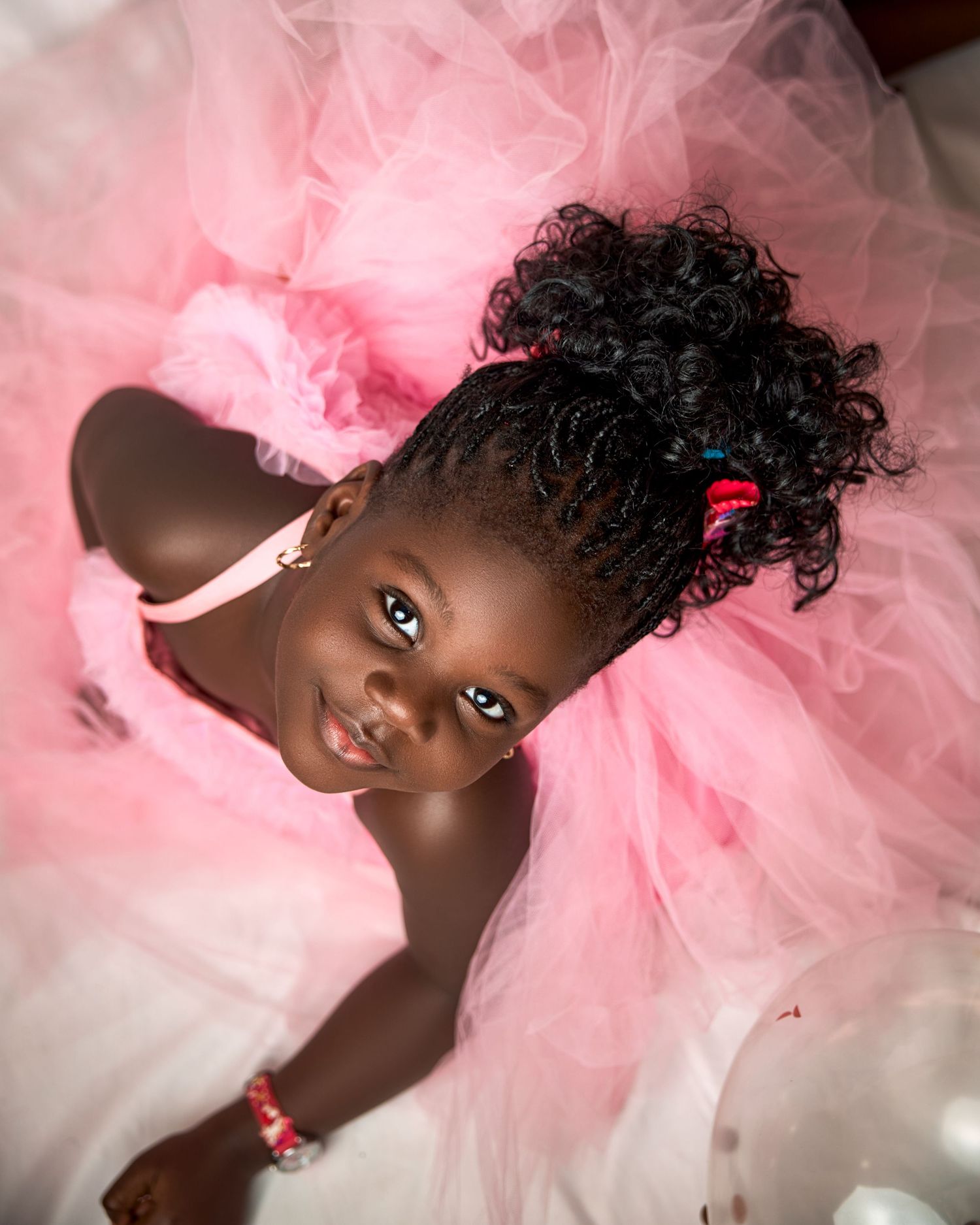 Young girl in pink tutu dress with curly updo hairstyle smiling up at camera in a joyful portrait.