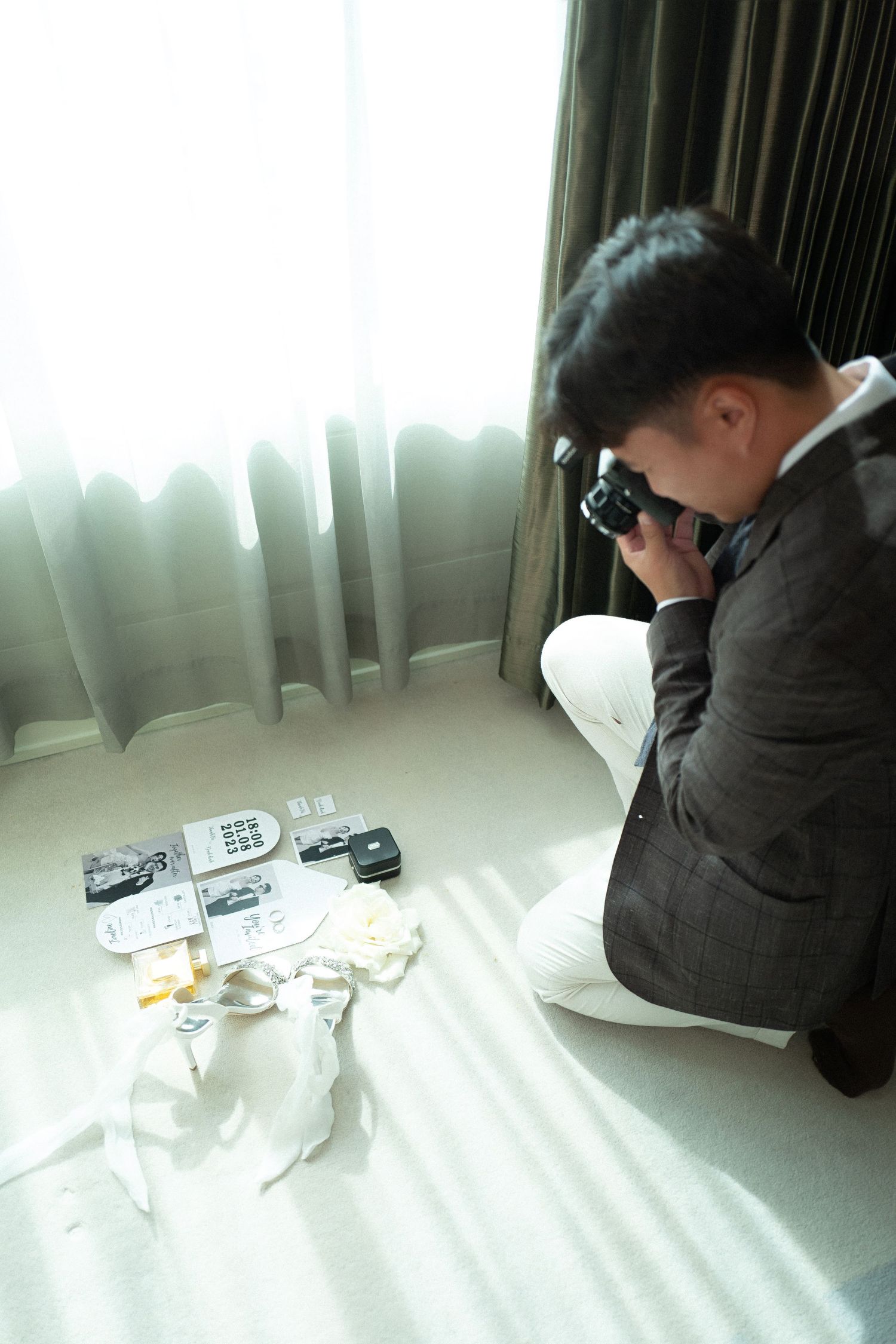 Groom in suit photographing wedding details arranged on hotel room floor near window.