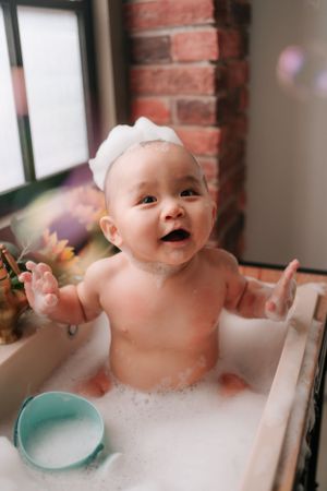 Happy baby enjoying bath time with bubbles in white tub against brick wall快樂的寶寶在白色浴缸裡，靠著磚牆，享受泡泡浴。