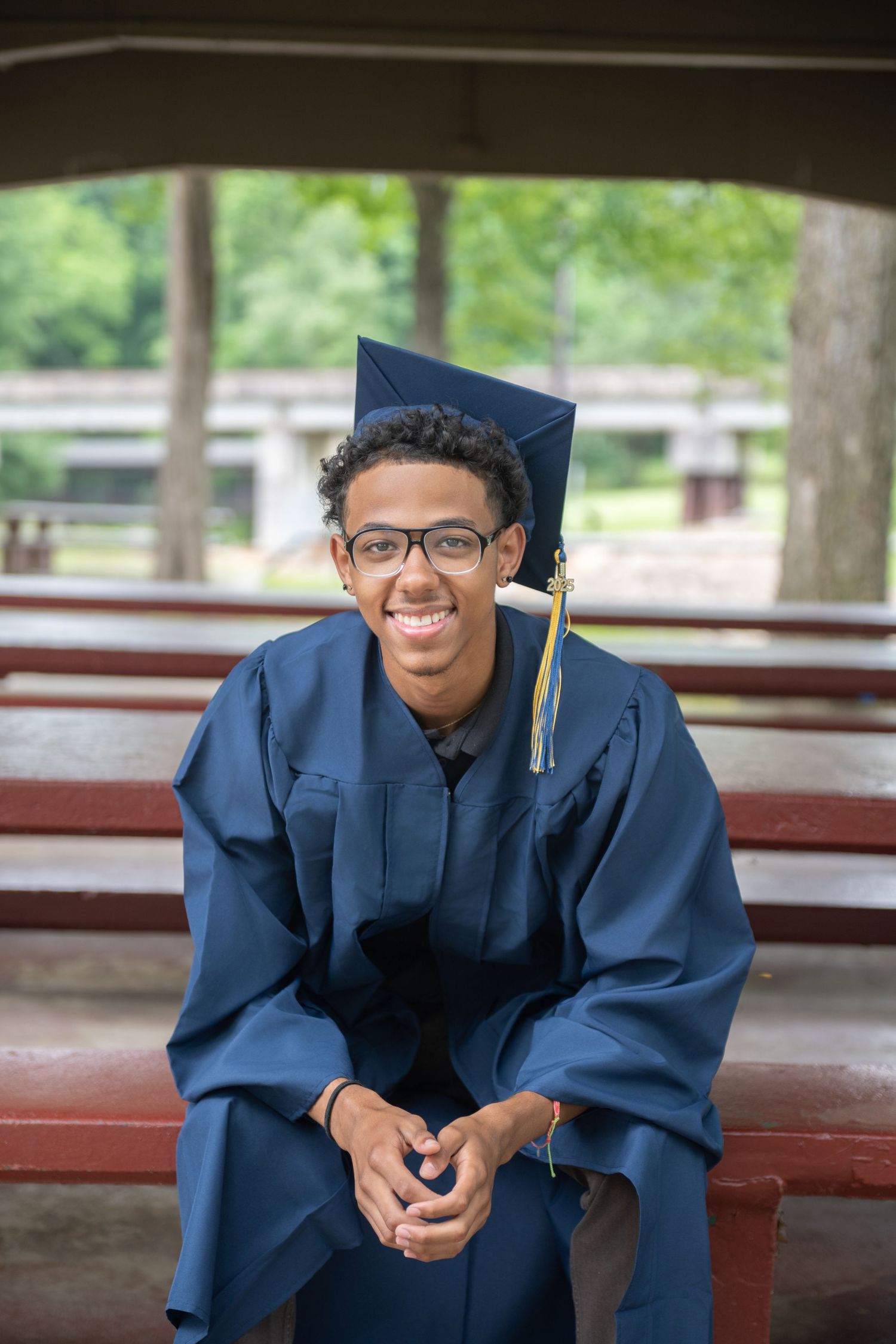 Smiling young man in navy blue graduation cap and gown seated on a bench outdoors, wearing glasses.