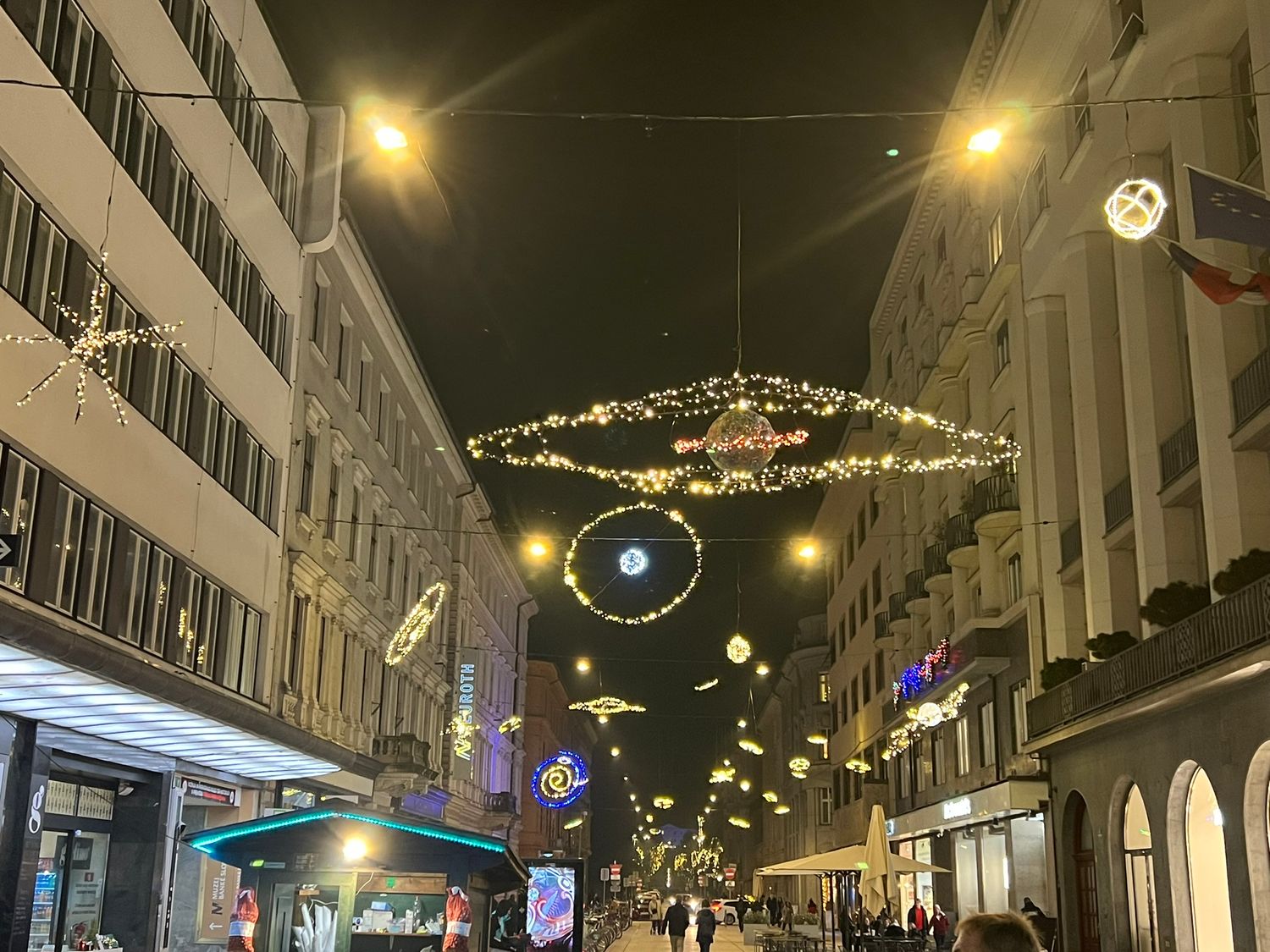 Festive street decorated with glowing chandelier lights and holiday ornaments illuminating a European shopping street at night.