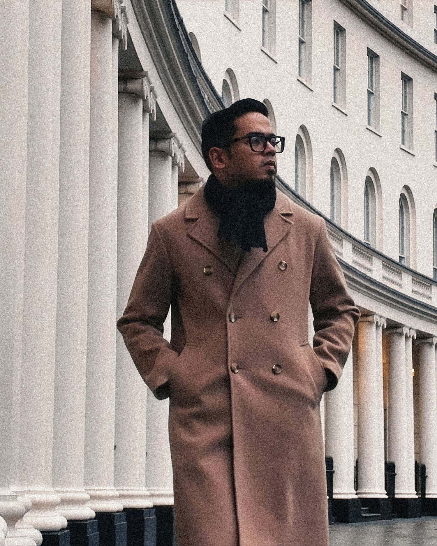 Man in camel overcoat and scarf standing by white columns of a classical building.