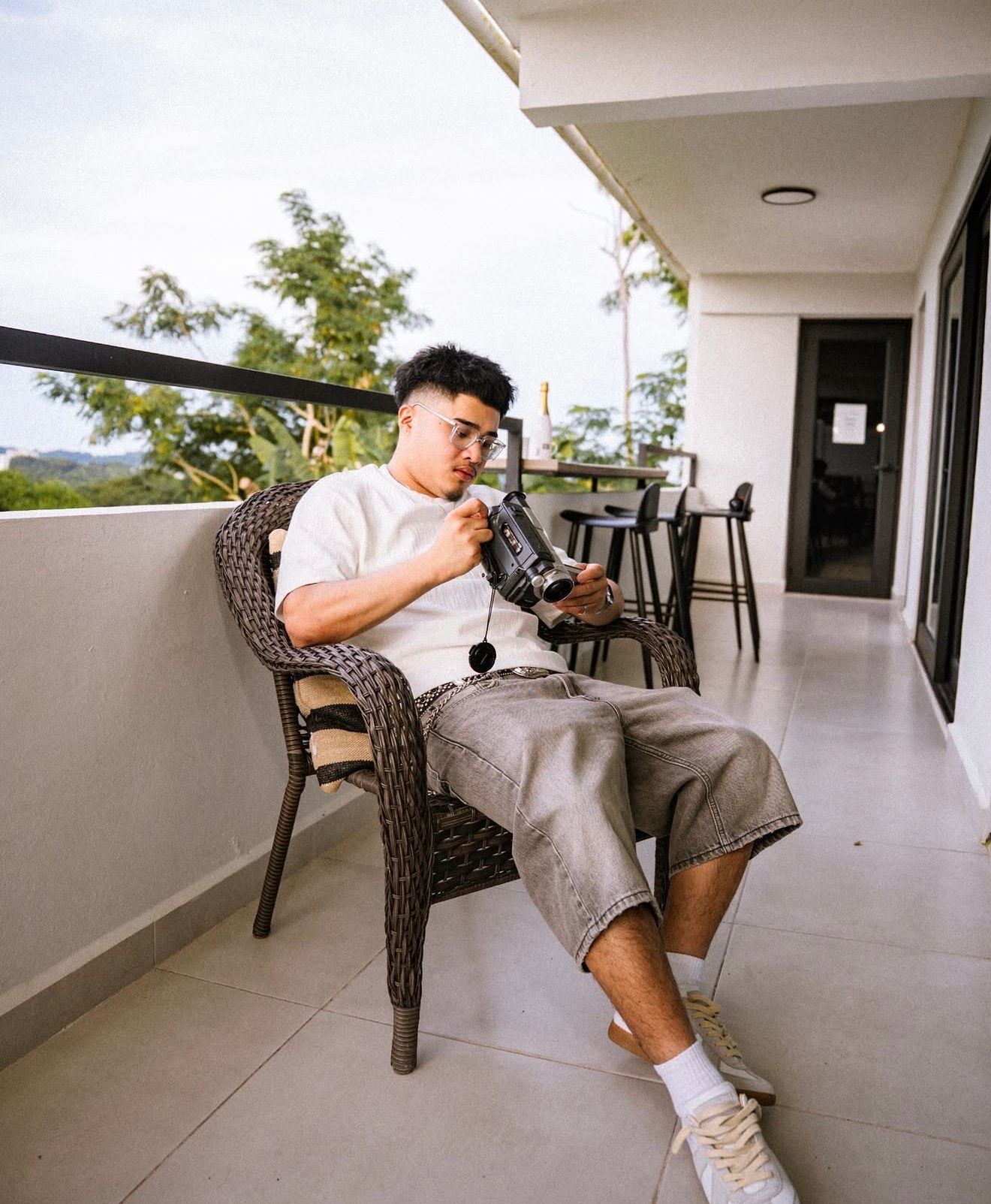 Young man sitting on balcony chair examining a camera with scenic outdoor view in background.