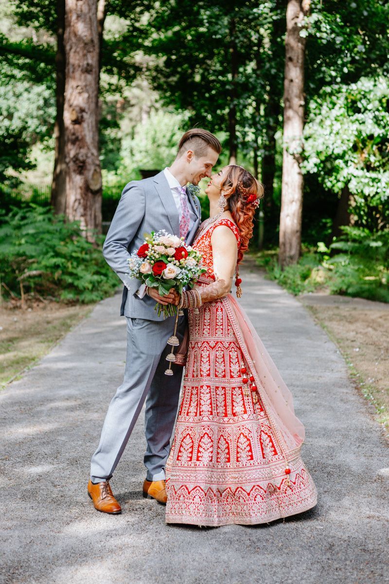 A bride in a red Indian lehenga and groom in gray suit share a romantic moment on a tree-lined path at the pumping house at ollerton