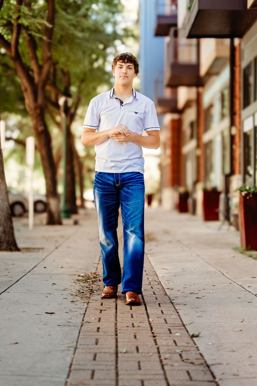 Young man in white polo shirt and blue jeans walking confidently on a city sidewalk lined with trees in Keller, TX.