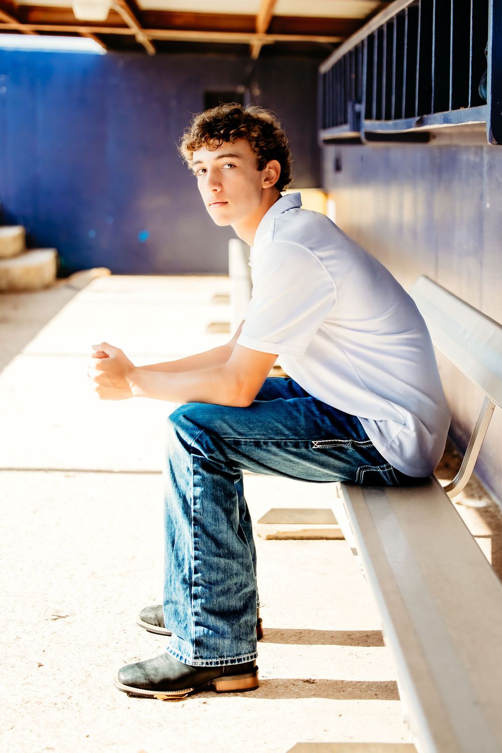 Teen boy in white shirt and jeans sitting on bleachers in a dugout during a sunny day in Keller, TX.