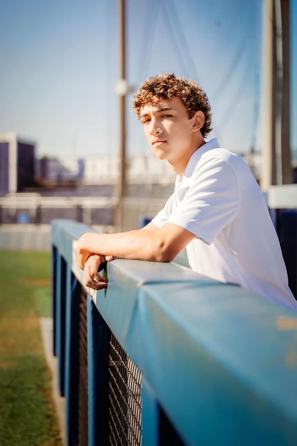 Young man with curly hair in white t-shirt leaning on blue railing at stadium on sunny day in Keller, TX.