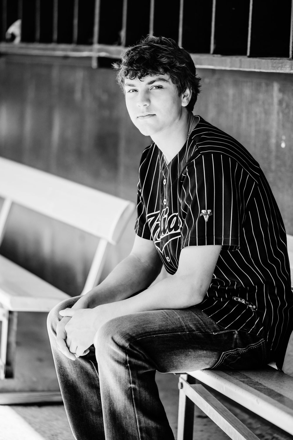 Black and white portrait of a teenage boy in a baseball jersey sitting in a dugout bench, looking at the camera in Keller, TX.