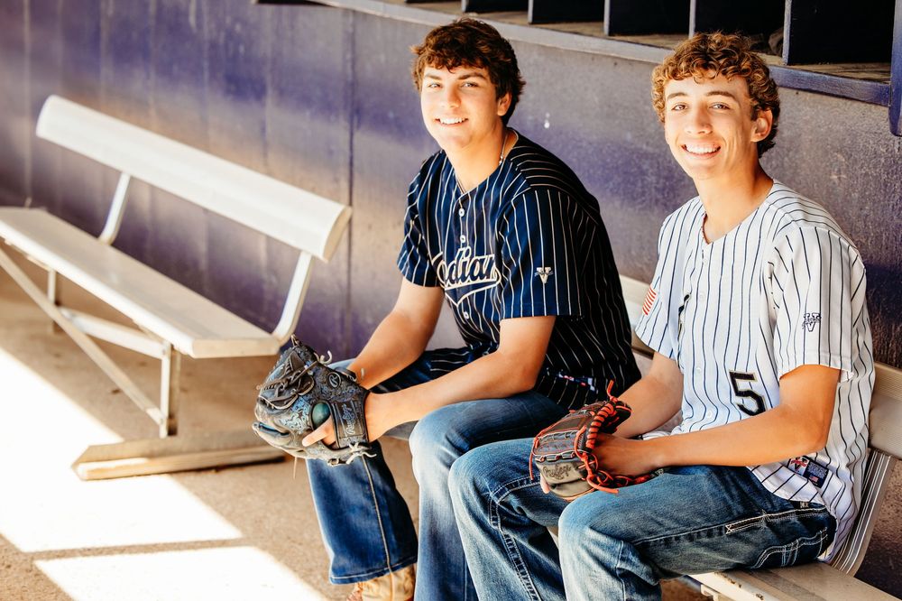 Two teenage baseball players in uniforms sit in dugout smiling for senior portraits in Keller, TX.