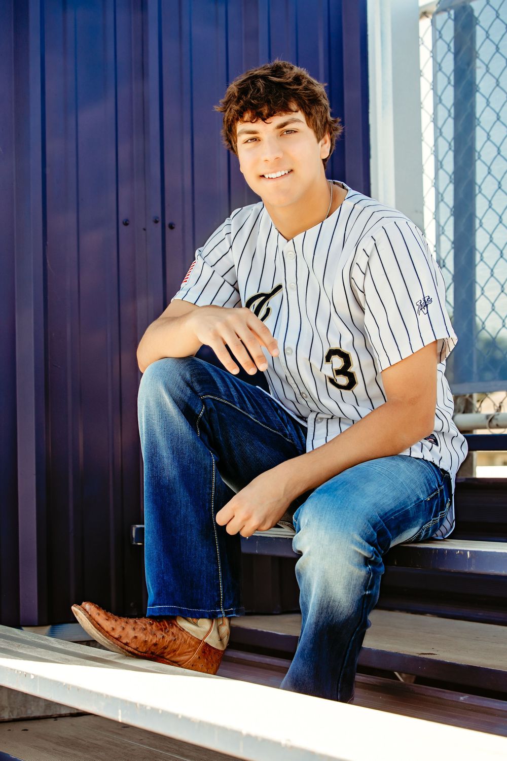 Young man in pinstripe baseball jersey number 3, jeans, and cowboy boots sitting on bleachers smiling in Keller, TX.