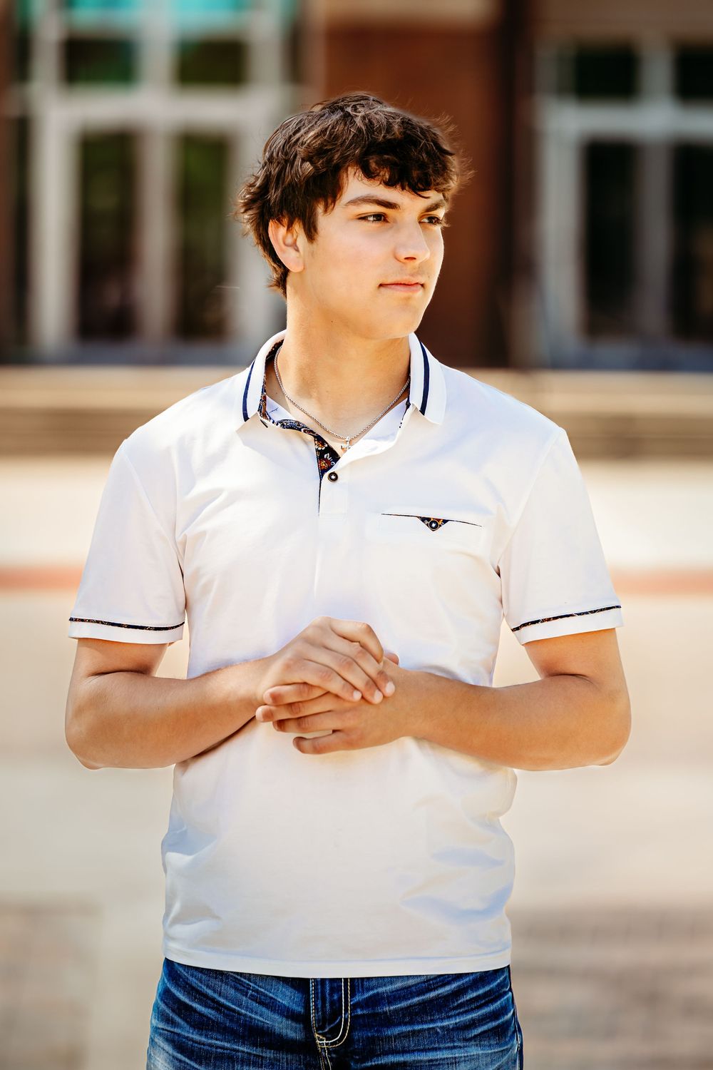 Young man in white polo shirt and jeans stands outdoors, hands clasped, with a building in the background in Keller, TX.