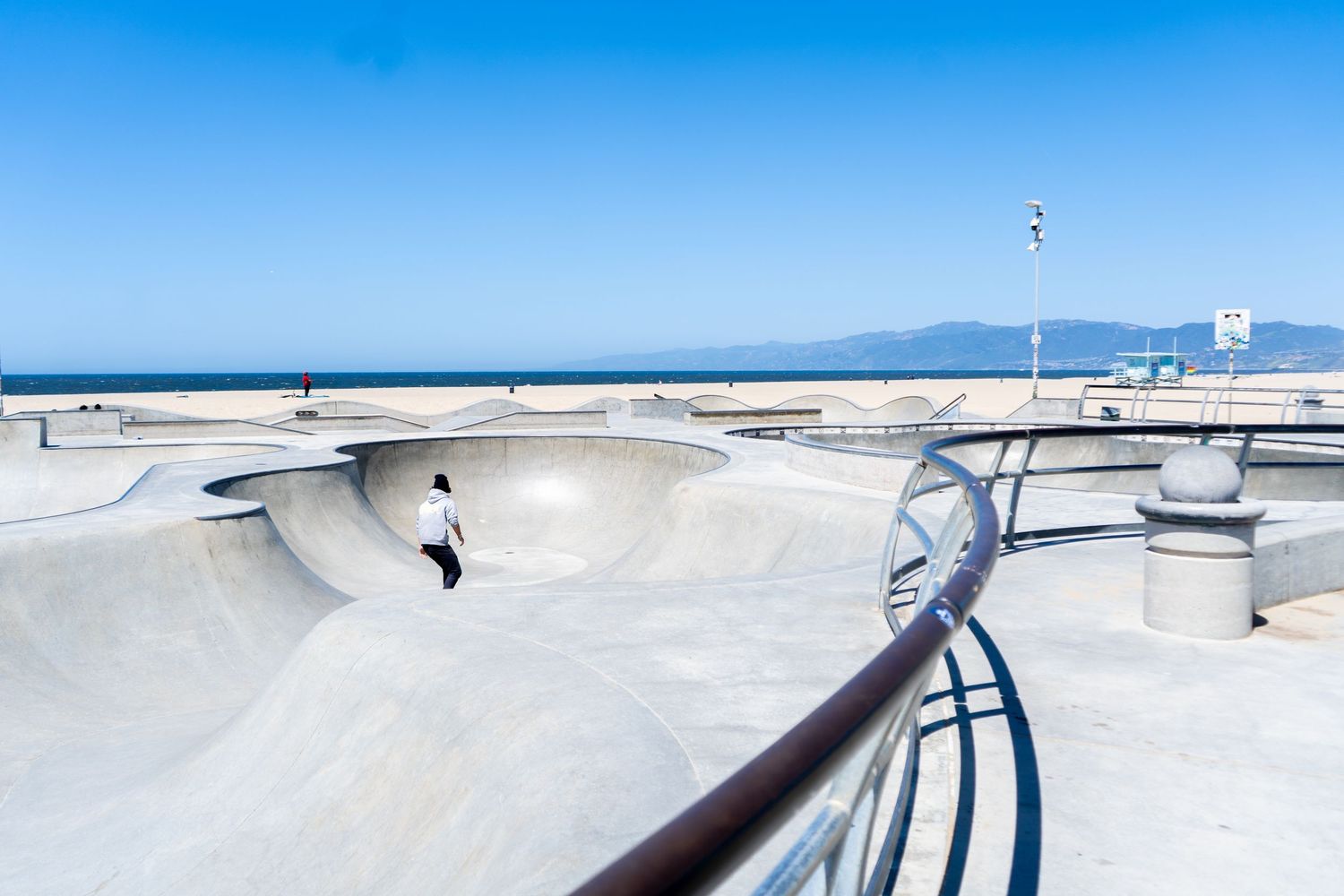 A skateboarder performs tricks at a concrete skate park with ramps and rails under bright blue skies.