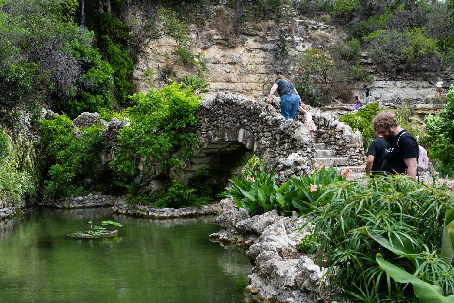 Two visitors explore a rustic stone bridge over a serene pond in a lush garden with limestone cliffs.