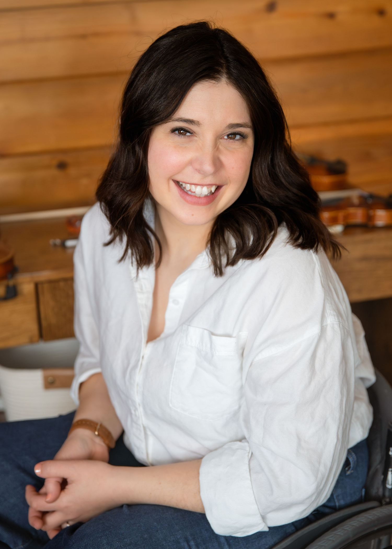 Smiling young woman with dark hair wearing a white button-up shirt, seated in a warmly lit wooden studio setting.