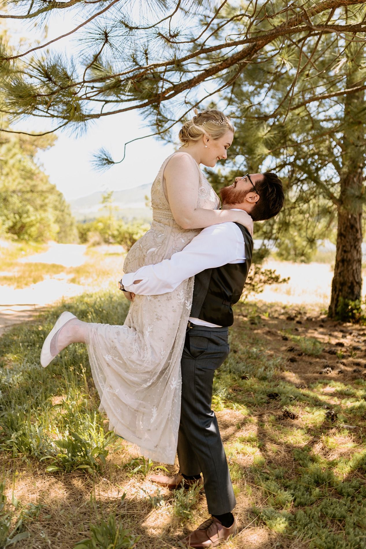 Groom lifts bride in wedding attire for a romantic kiss outdoors under pine trees in a sunlit forest setting at Lake Cuyamaca, Julian.