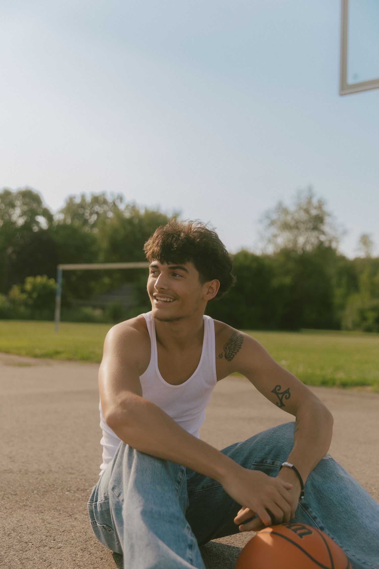 Young man with tattoos sitting on basketball court holding a ball, smiling outdoors on a sunny day.