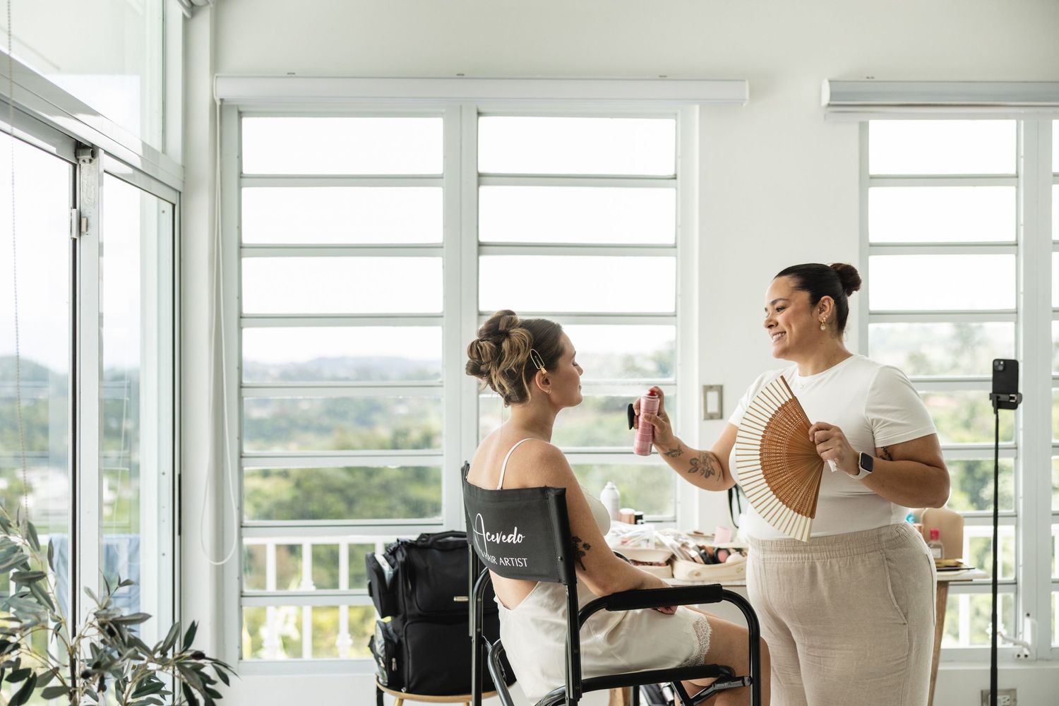 Makeup artist applies bridal beauty touches to a bride seated by bright windows.