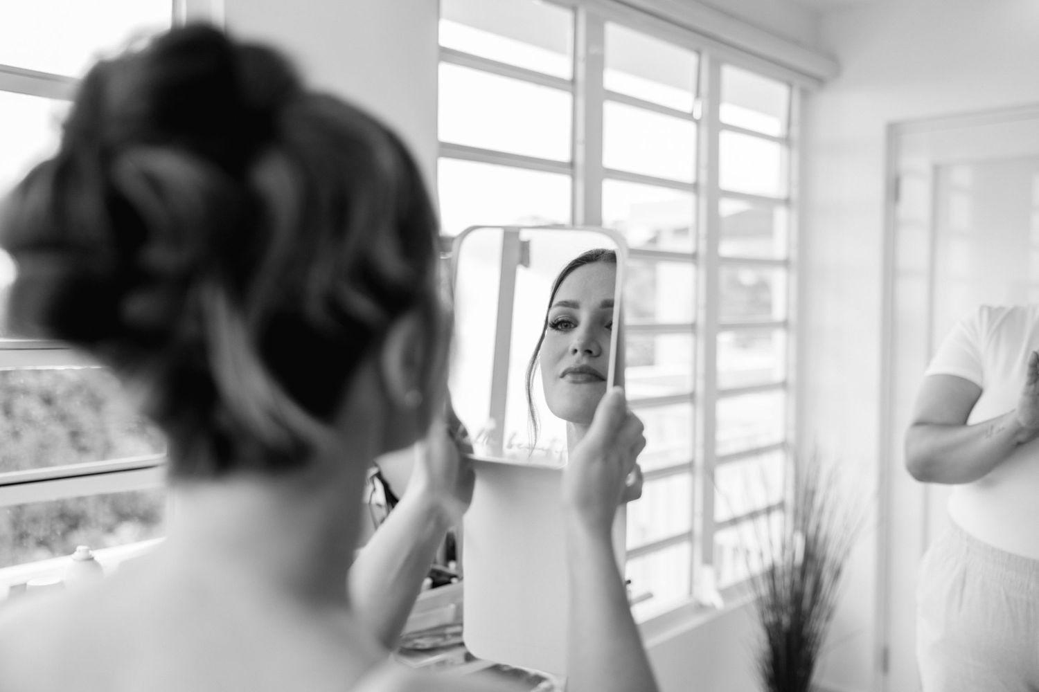 Black and white photo of a bride examining her makeup in a handheld mirror while getting ready.