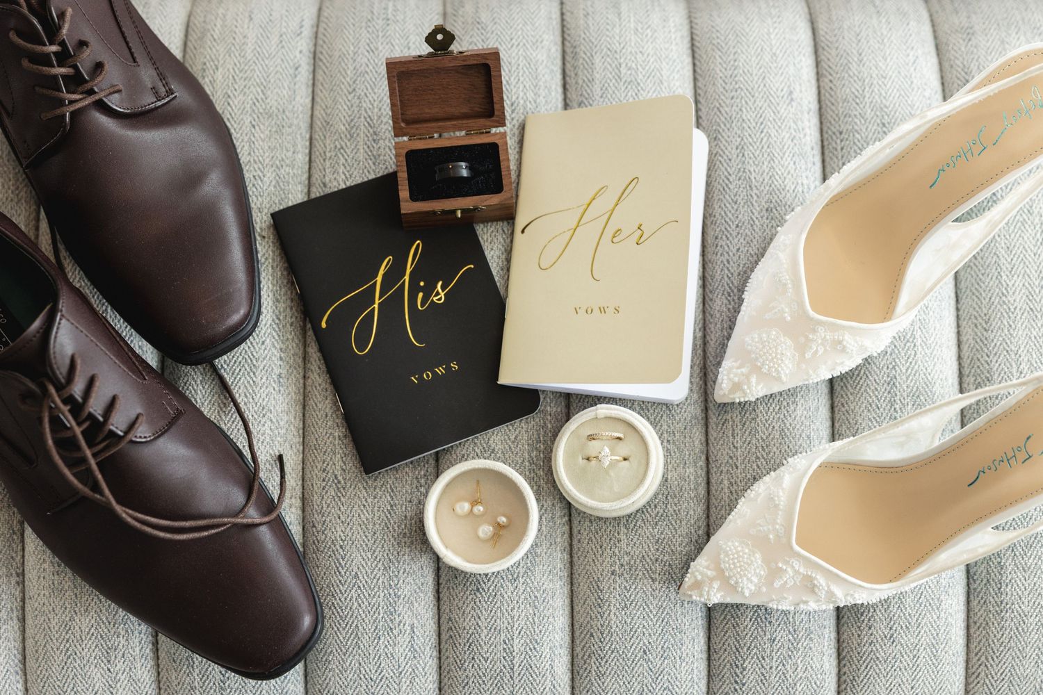 Flat lay of wedding accessories including brown dress shoes, lace heels, vow books, and rings.