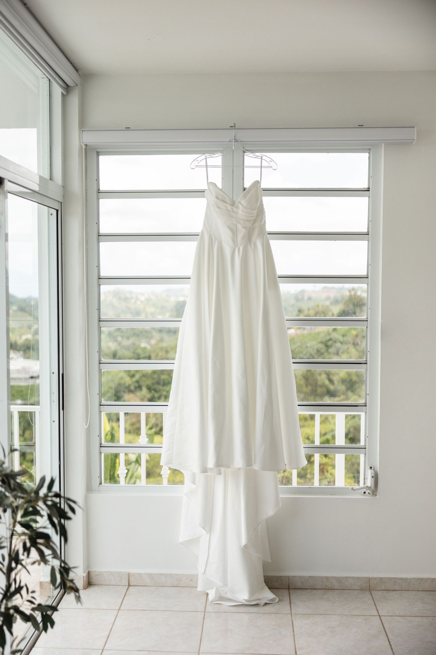 White wedding dress hanging on a hanger in front of a bright window with louvered shutters.