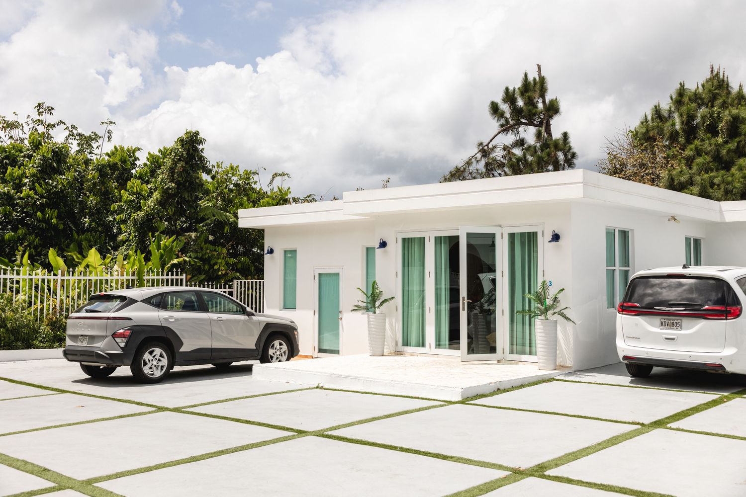 Modern white mid-century home exterior with two cars parked in a spacious driveway surrounded by tropical greenery.