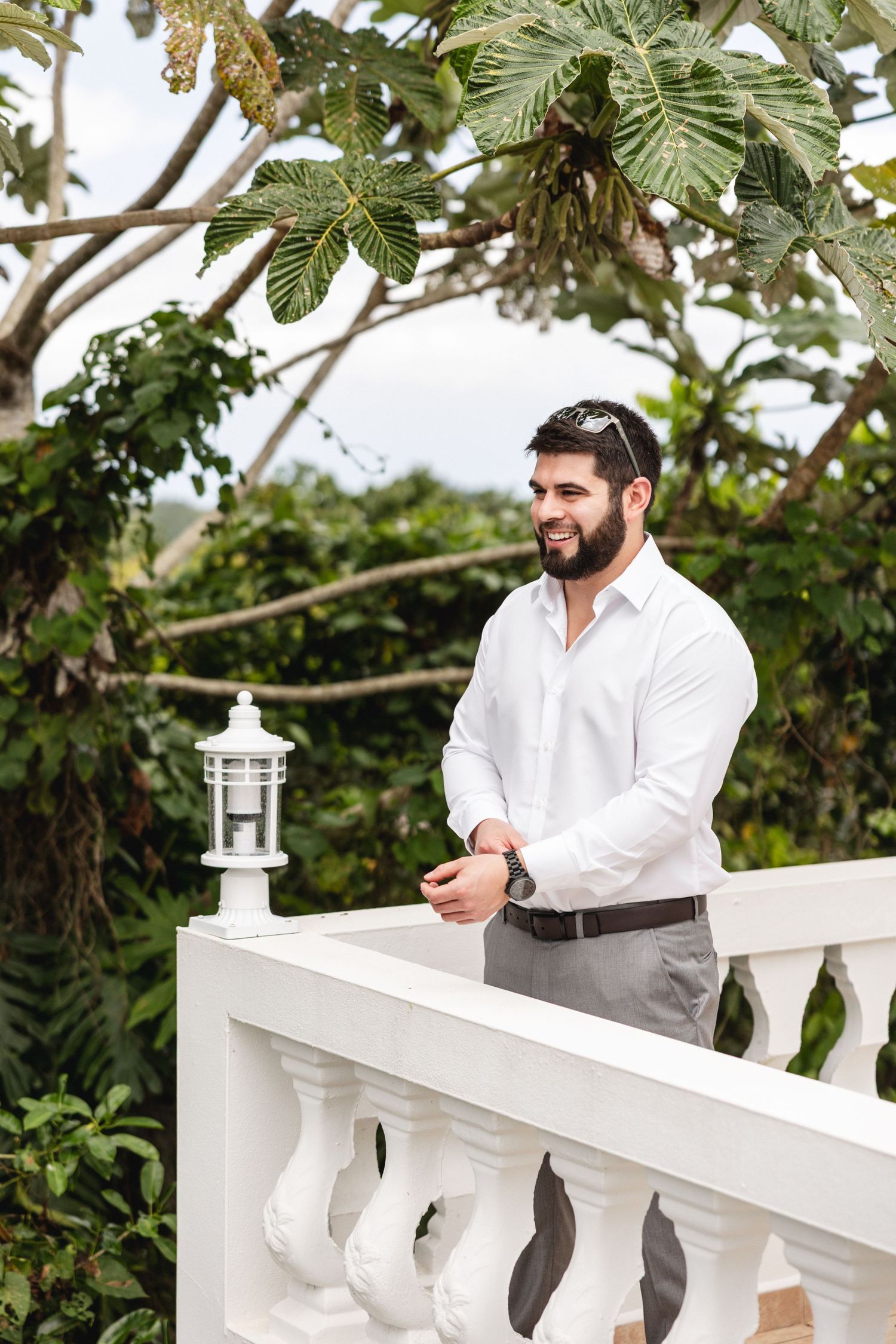 Handsome bearded groom in white shirt smiling on a white balcony surrounded by tropical greenery.