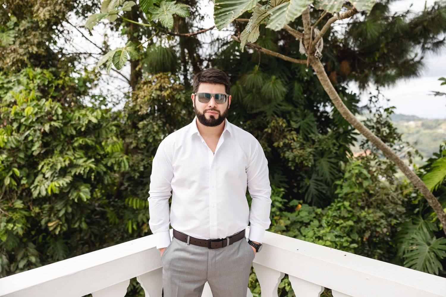 Stylish man in white shirt and sunglasses standing on white balcony with tropical greenery.