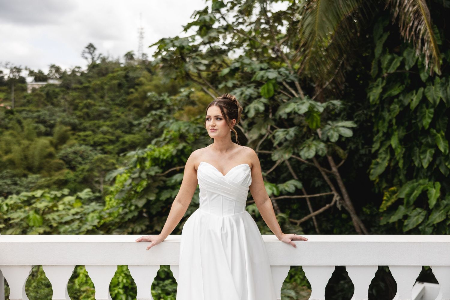 Elegant bride in a white strapless gown standing on a balcony with lush green trees behind her.
