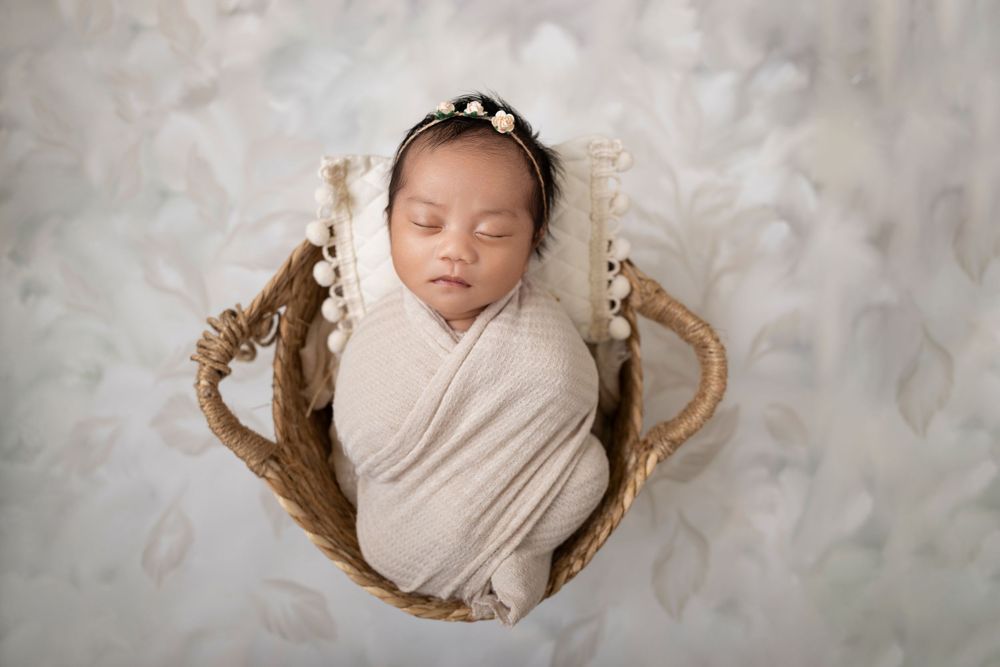 Newborn baby swaddled in cream wrap sleeping peacefully in a wicker basket with floral headband in Newborn Studio Photography in NJ.