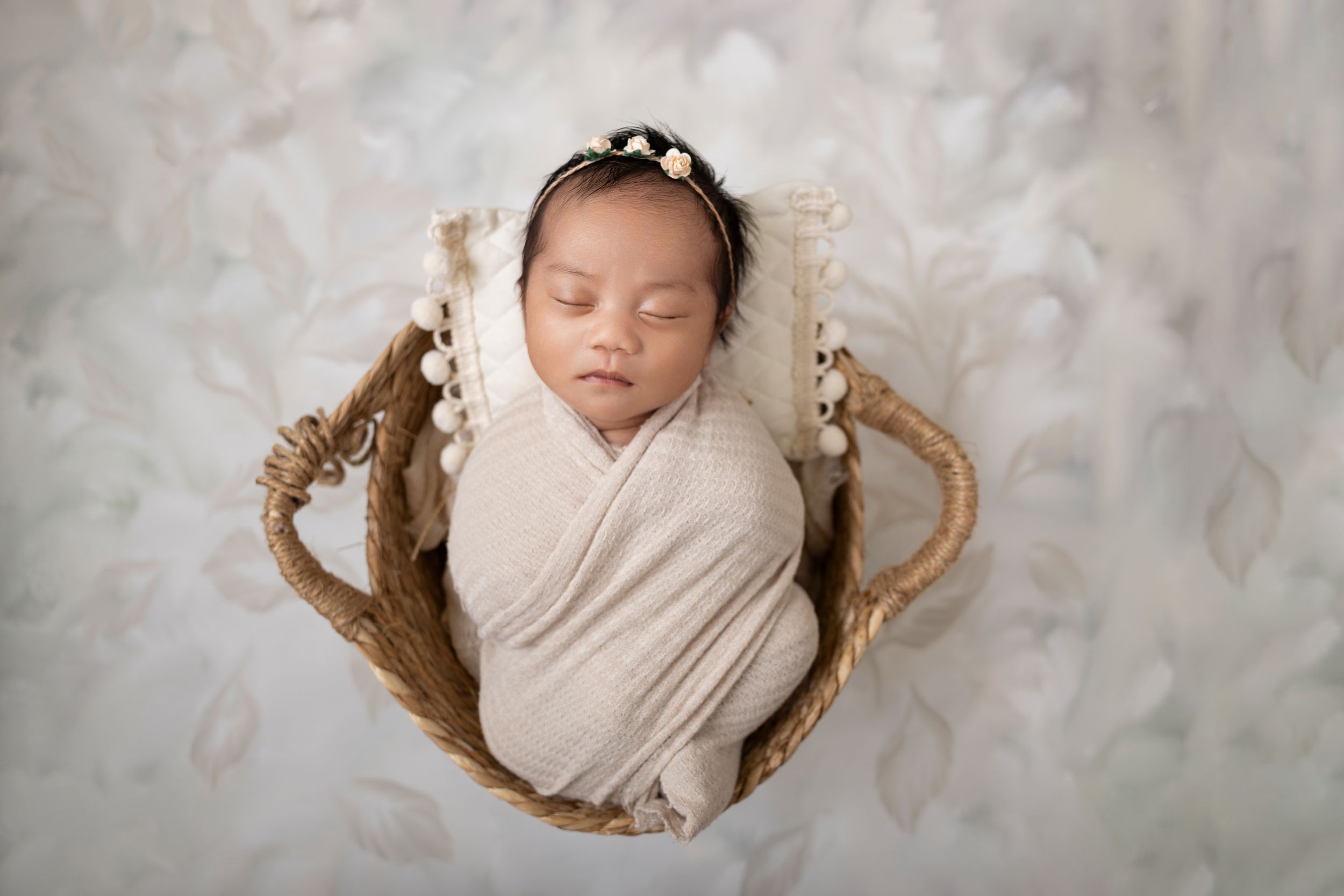 Newborn baby swaddled in cream wrap sleeping peacefully in a wicker basket with floral headband in Newborn Studio Photography in NJ.
