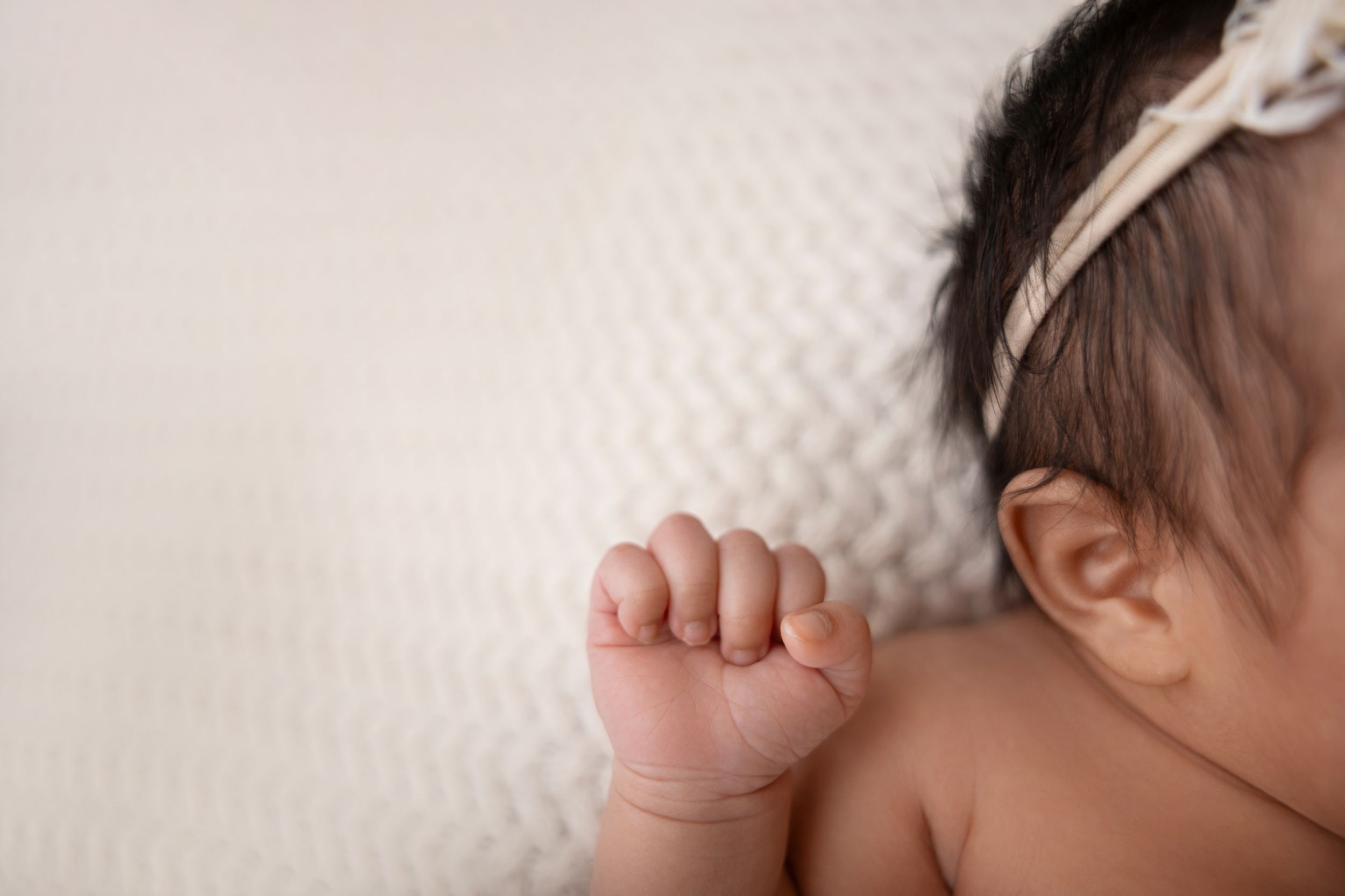Close-up of a newborn baby's tiny fist and ear, wearing a white bow headband, resting on a soft white blanket in NJ photography studio