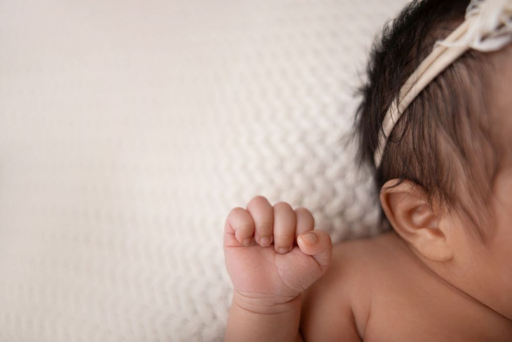 Close-up of a newborn baby's tiny fist and ear, wearing a white bow headband, resting on a soft white blanket in NJ photography studio