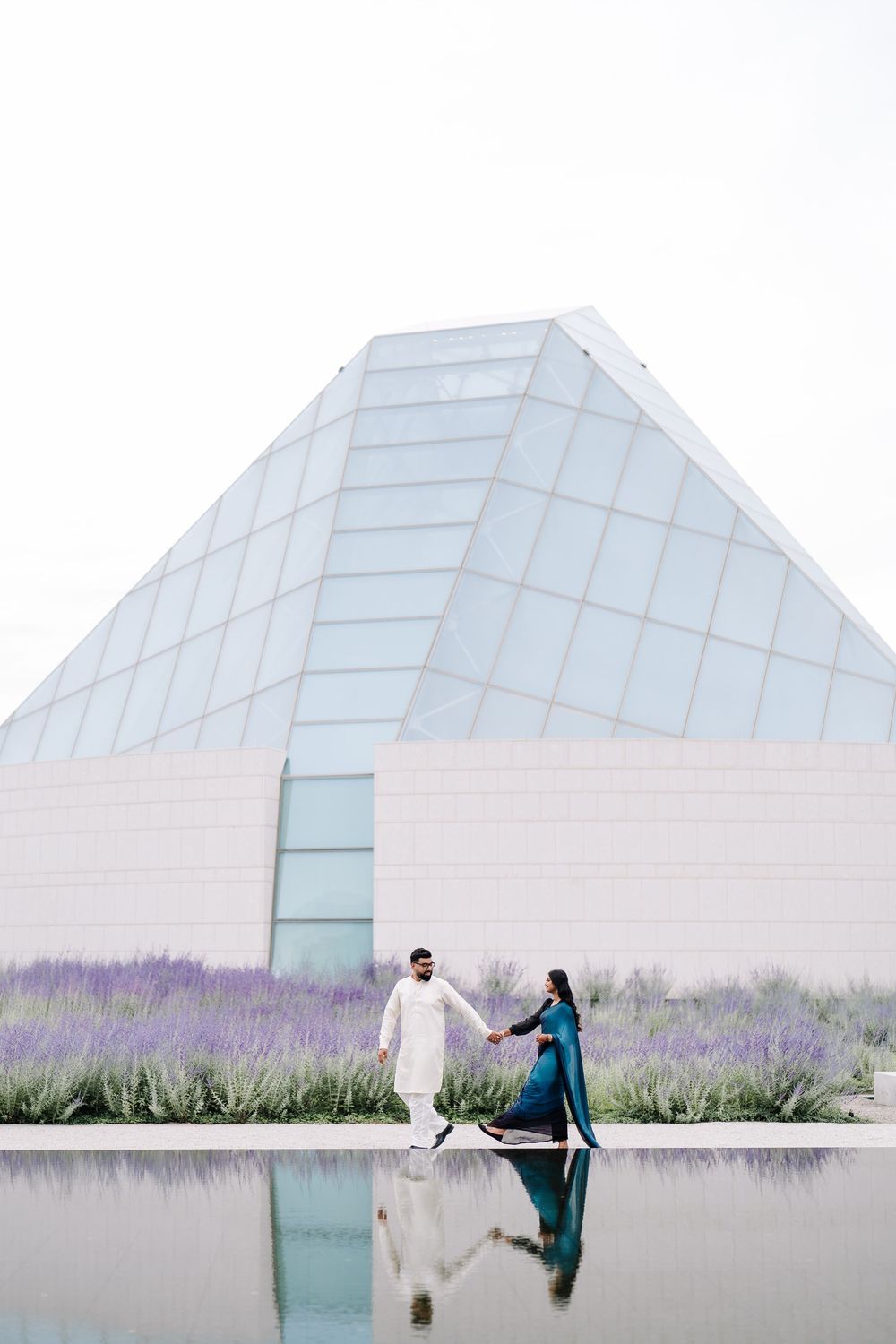 Couple in traditional South Asian attire holding hands by a reflective pool near a modern glass pyramid building with lavender.