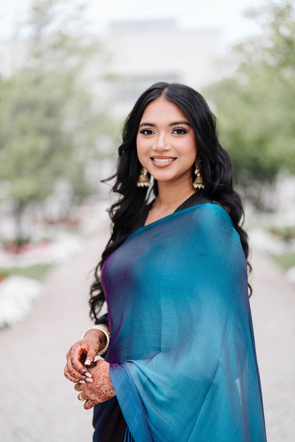 A young woman in a teal blue saree with gold jhumka earrings and henna hands smiles outdoors.
