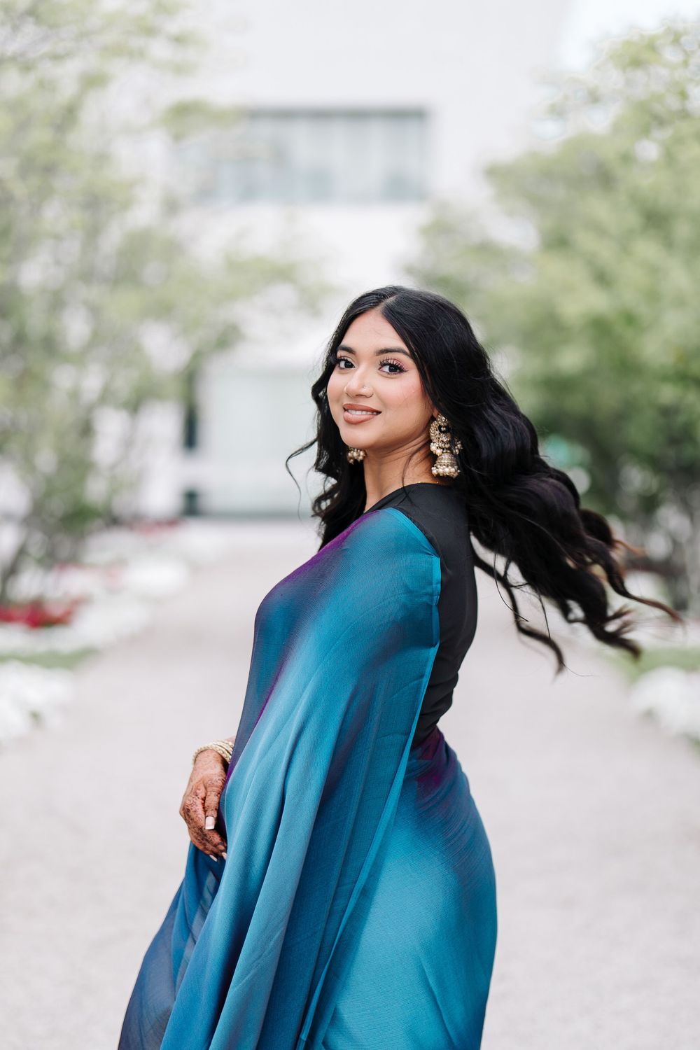 A young woman in a teal sari with gold earrings poses outdoors, her black hair flowing gracefully.