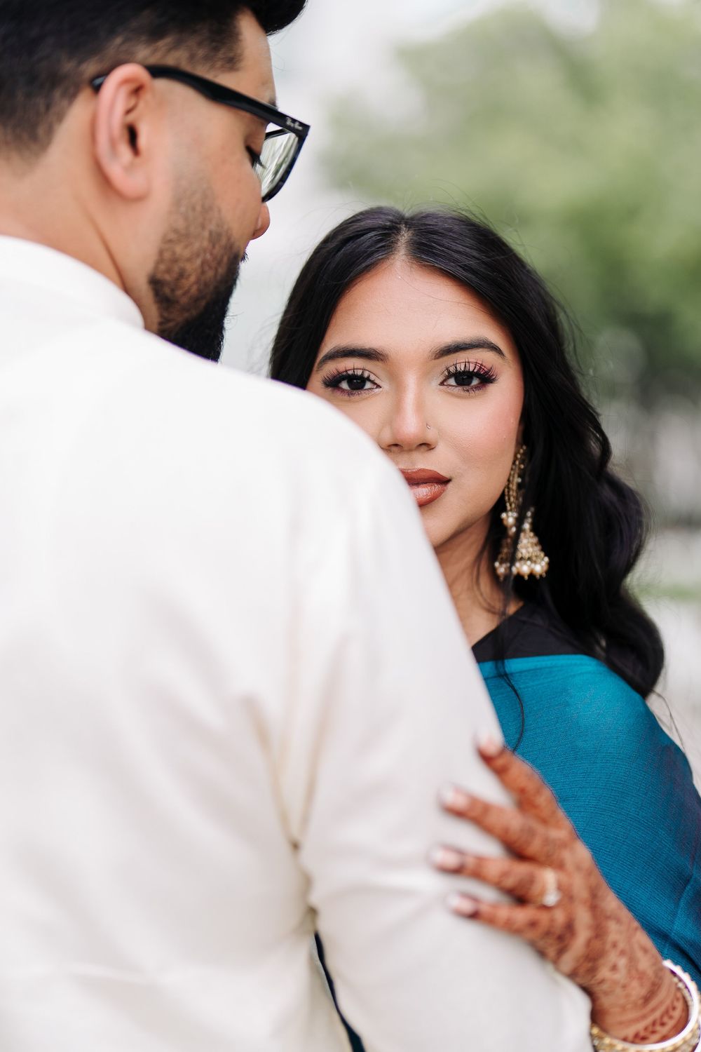 A woman in a teal dress with gold jewelry and henna gazes at a man in a white suit during an engagement shoot.