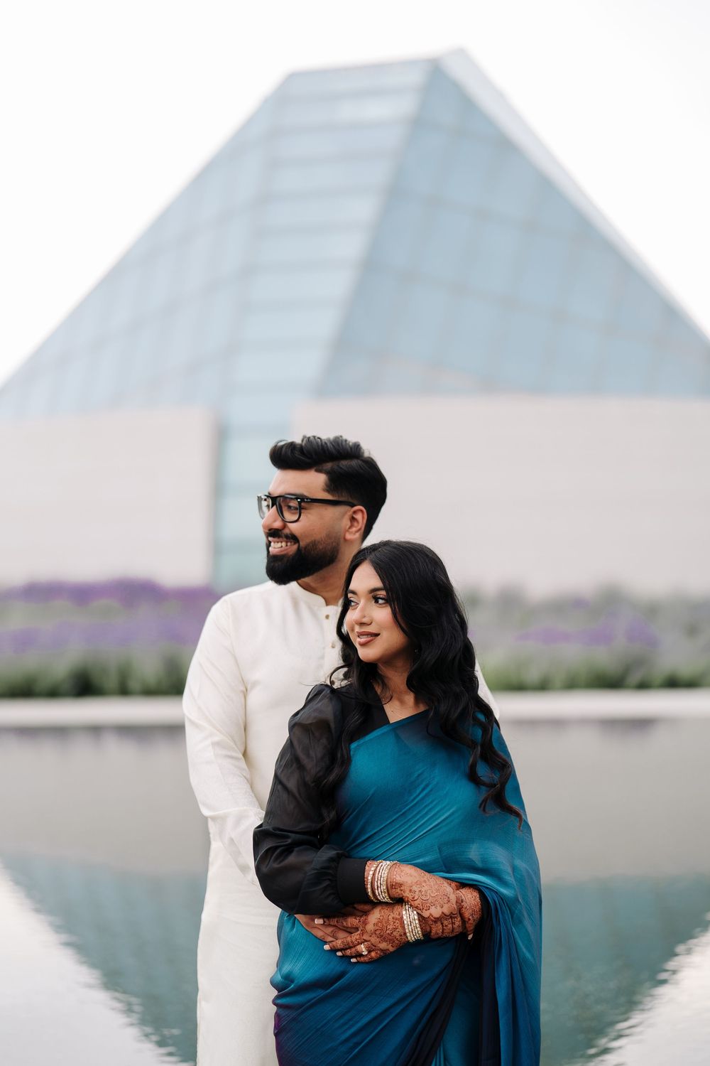 Couple in traditional South Asian attire posing near a reflective pool with a glass pyramid building backdrop.